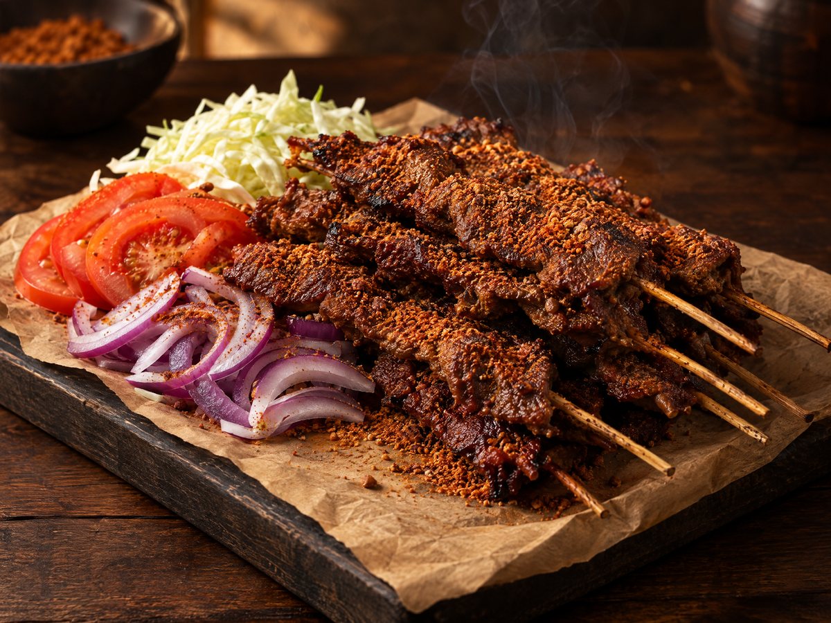 Spice-crusted suya beef skewers on paper with sliced onion, tomato and cabbage